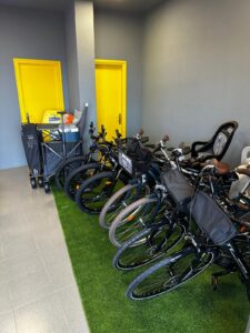 Bicycle storage room at KUDO Rent with multiple bikes, child seat, and storage carts on green artificial grass near yellow doors, ideal for bike rentals and outdoor activity convenience.