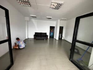 Spacious commercial interior with white walls, tiled flooring, and modern ceiling lights, featuring a black sofa and an individual working on a small project, ideal for office or business rental with KUDO Rent.
