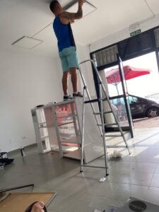 High-angle shot of a man installing a ceiling light fixture using a ladder in a commercial space, with a large glass door and outdoor seating visible in the background.