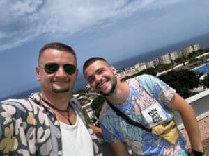 Vibrant outdoor photo of two smiling men enjoying a sunny day, wearing casual summer shirts, with scenic ocean and cityscape in the background, ideal for travel and vacation content.