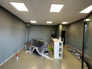 Modern office interior renovation in progress, featuring freshly painted walls, ceiling lighting, a partially covered sofa, cleaning supplies, and construction tools on the floor.
