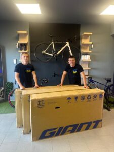 High-quality image of two men from KUDO Rent smiling behind large Giant bicycle box, with a mounted bike on the wall and bicycle accessories, in a modern showroom for bike rentals and sales.