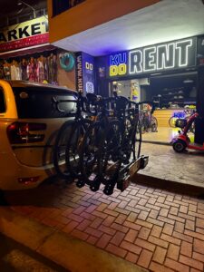 Bike rack mounted on the back of a yellow car outside KUDO Rent store at night, with bicycles for rental and illuminated sign in the background.