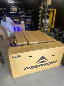 Boxes of Merida bicycles inside a bike shop, ready for delivery or display, with bike accessories like helmets and helmets in the background, highlighting bicycle rental and sales services.