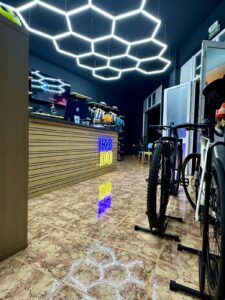 Bike rental shop interior with illuminated ceiling lights, display of cycling helmets, bikes, and accessories, modern decor, wooden counter, and neon KUDO rent sign, showcasing urban bike rental services.