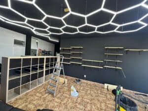 Modern interior space with black accent wall featuring mounted wooden shelves, contemporary honeycomb ceiling lighting, and a mix of unfinished furniture and tools, indicating ongoing setup or renovation.
